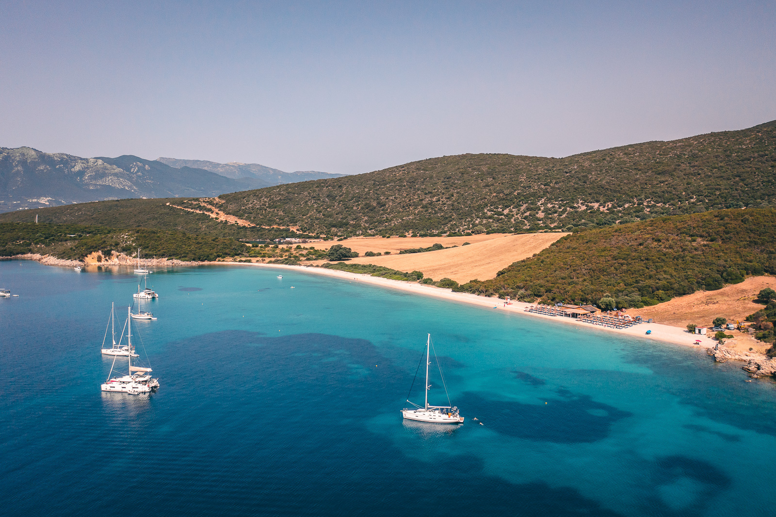 Aerial view of Vathiavali bay, a long crescent of pale sand beside golden fields, with turquoise shallows and sailing boats anchored offshore