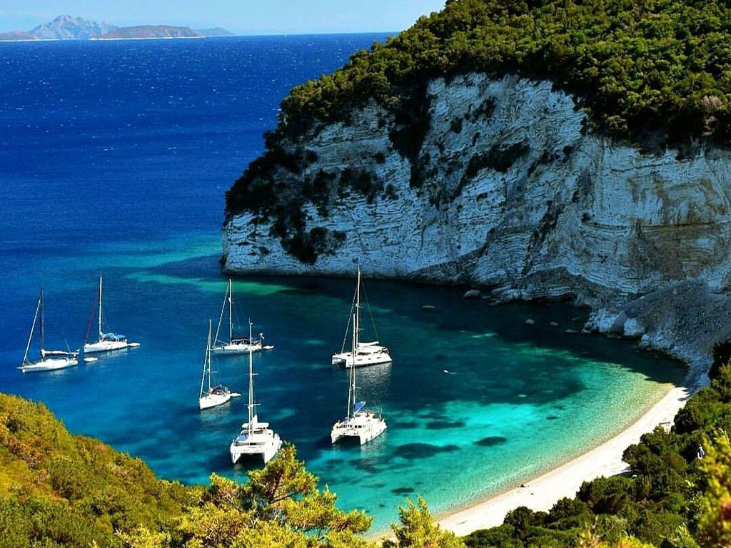 Aerial view of white cliffs, a small beach and boats anchored in a turquoise cove at Atokos