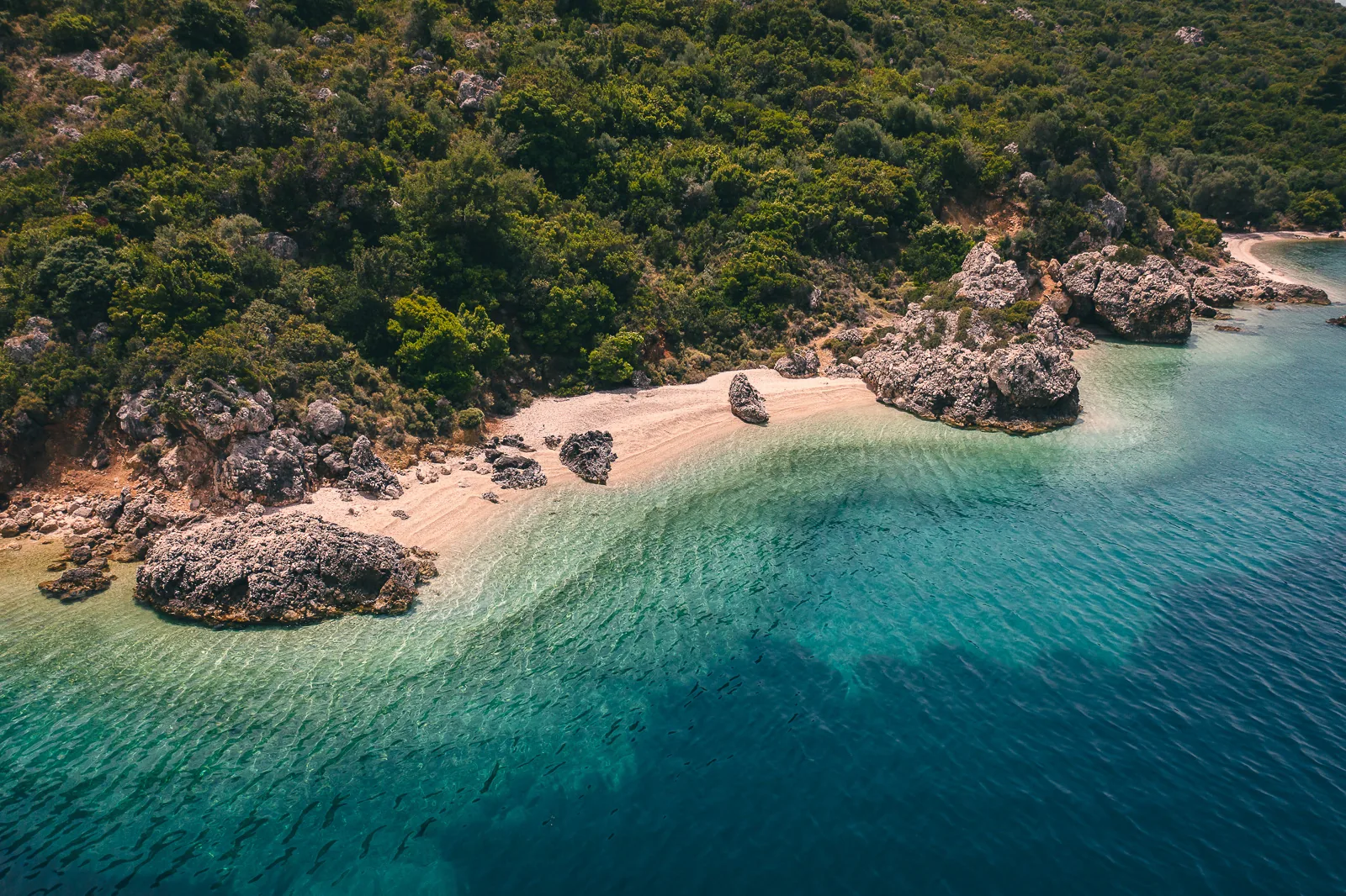Aerial view of a secluded cove with pale sand, turquoise water and a forested hillside near Paleros
