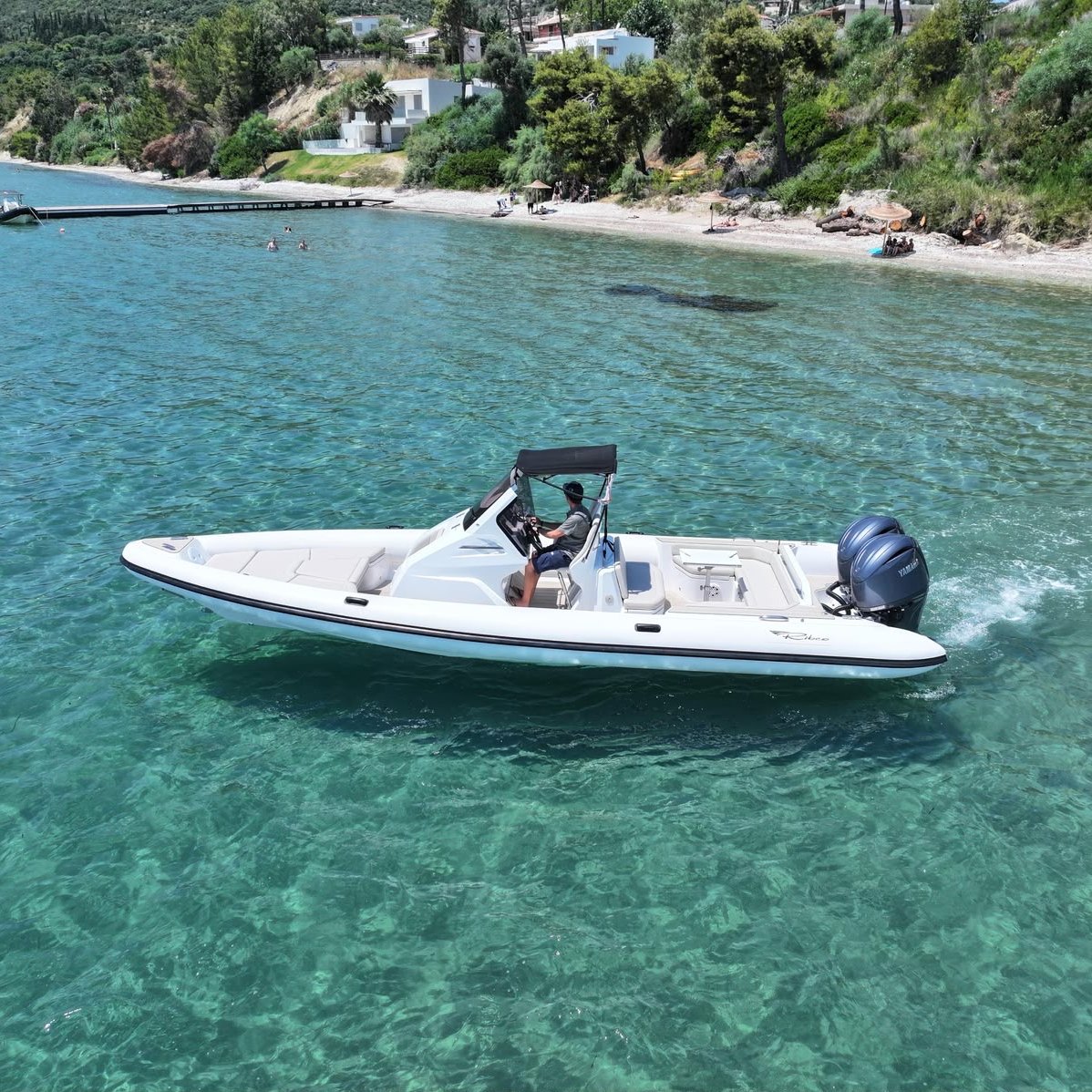 White rigid-inflatable boat with a canopy and twin outboard motors on clear turquoise water off a pebble beach
