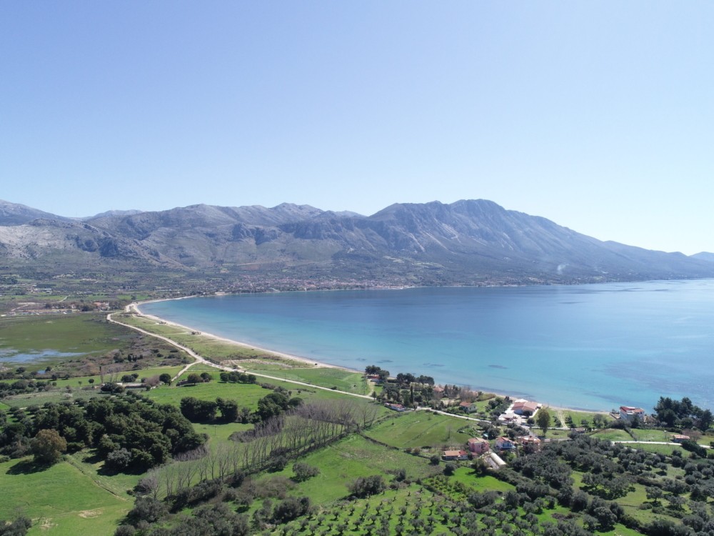 Aerial view of Potamaki beach, a long sandy bay bordered by green fields with mountains across the water