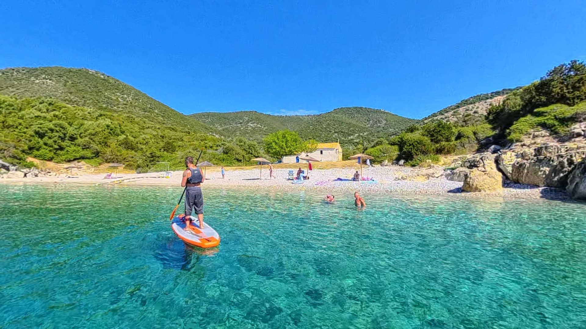 Clear turquoise water off a pebble beach at Ai Giannis, with a small stone church behind the shore, green hills rising behind and a paddleboarder on the water